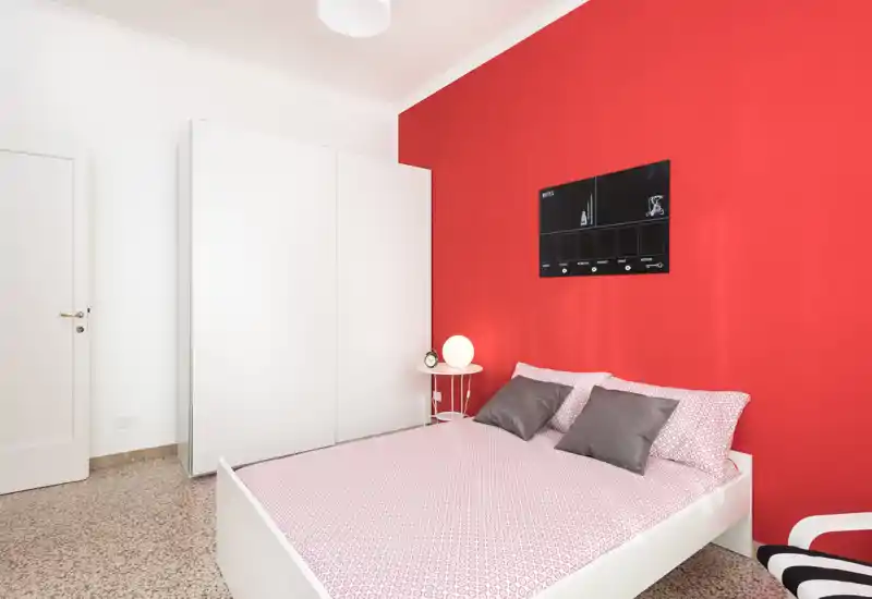 Camera da letto moderna e luminosa con letto matrimoniale davanti a una parete rossa vivace, armadio bianco e lampada da comodino che creano un ambiente pulito e accogliente.