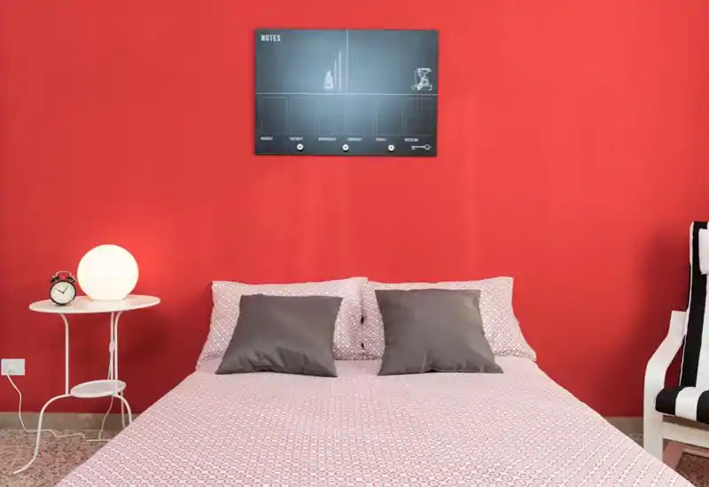Camera da letto luminosa e curata con letto matrimoniale centrale su una parete rossa vivace, due cuscini grigi e biancheria coordinata; comodino con lampada e sveglia per un tocco accogliente.