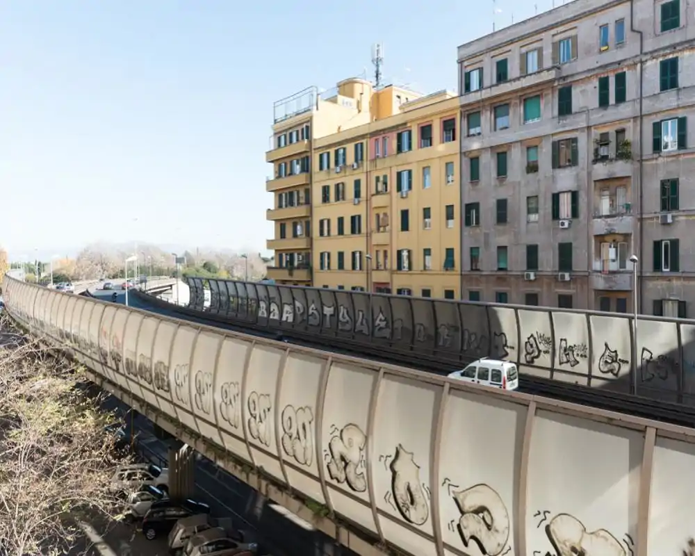 Veduta esterna urbana di una strada sopraelevata con pannelli protettivi coperti di graffiti, edifici residenziali adiacenti e cielo sereno.