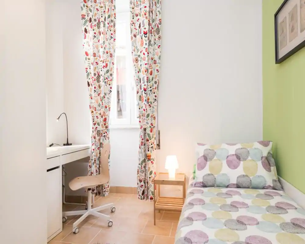 Habitación individual luminosa y compacta con edredón estampado, mesita de noche y escritorio junto a la ventana. Las cortinas florales y la luz cálida crean un ambiente acogedor.