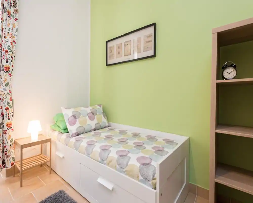 Habitación individual ordenada con cama blanca y ropa de cama estampada, lámpara de noche, cortinas coloridas y una pequeña estantería; luminosa, limpia y acogedora para un huésped.