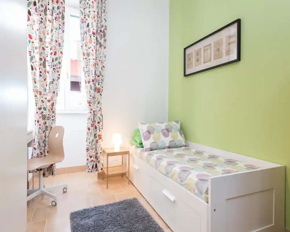 Dormitorio individual compacto con cama blanca tipo diván, ropa de cama estampada y alfombra suave; decoración luminosa y alegre con pared verde y cortinas florales que aportan luz natural.