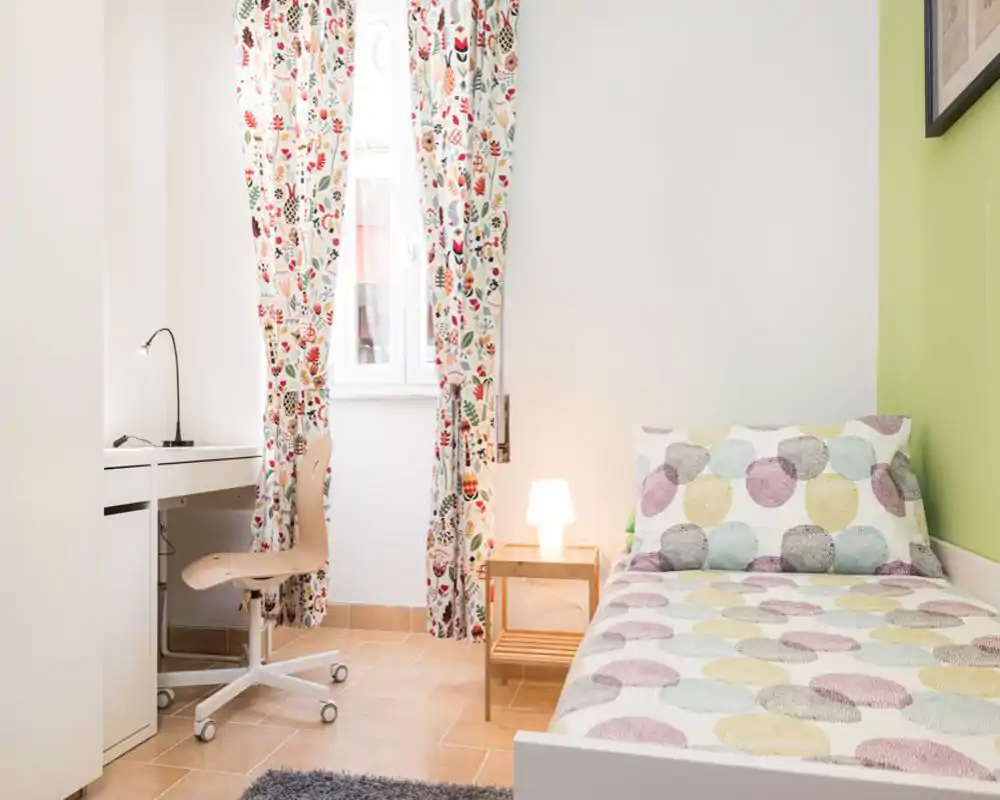 Dormitorio individual luminoso y bien presentado, con ropa de cama estampada, mesita con lámpara y un pequeño escritorio junto a la ventana — perfecto para un anuncio de alquiler acogedor.