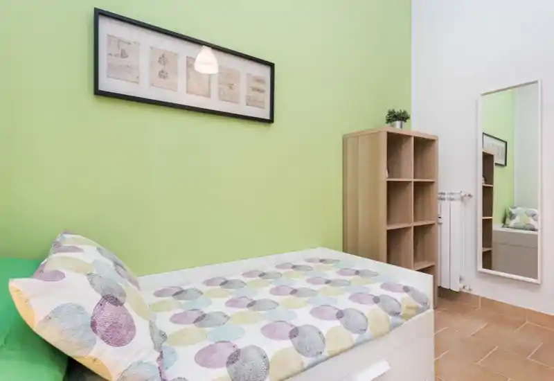 Dormitorio compacto y bien decorado con una cama individual con ropa de cama estampada, una estantería alta y un espejo de cuerpo entero. La pared verde claro y la luz suave crean un ambiente fresco y acogedor, perfecto para un anuncio de alquiler.