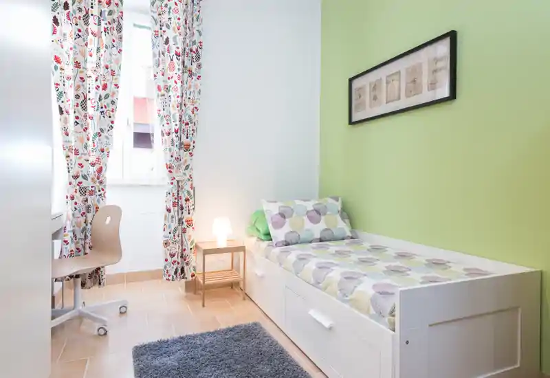 Habitación individual luminosa con una cama tipo sofá blanco, ropa de cama de lunares coloridos, cortinas estampadas y un pequeño escritorio junto a la ventana — ordenada, acogedora y bien iluminada.