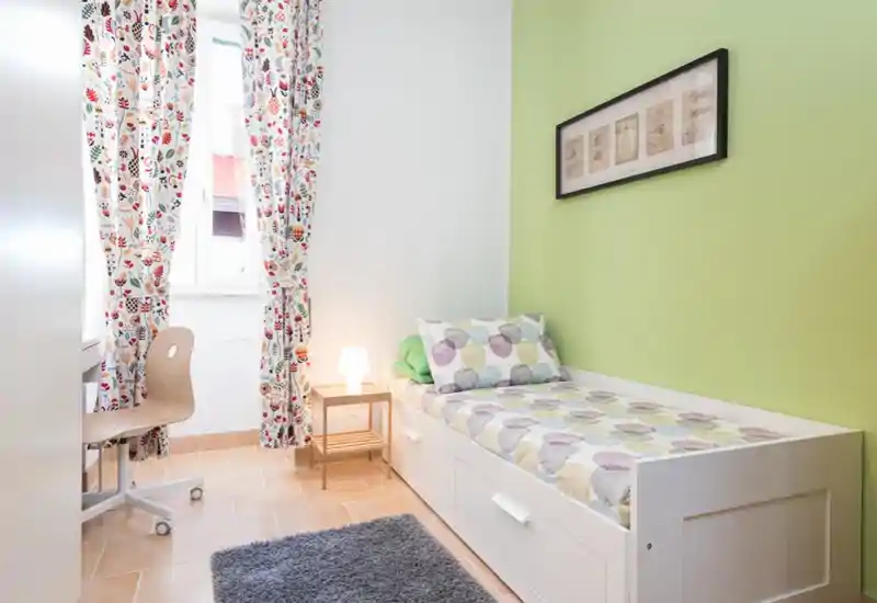 Camera da letto singola luminosa e ordinata con letto bianco, tende colorate a fantasia e un piccolo comodino — ideale per un affitto compatto o una stanza per gli ospiti.