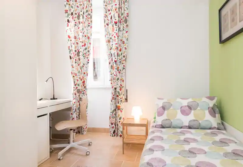 Dormitorio individual luminoso con cortinas estampadas y lámpara de mesita acogedora. La cama ordenada con ropa de cama colorida y un pequeño escritorio hacen el espacio práctico y agradable.