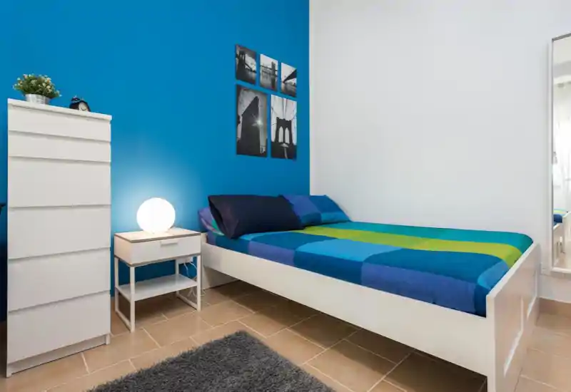 Camera da letto moderna e luminosa con struttura del letto bianca, biancheria dai toni blu-verde e un comodino con lampada soffusa. Arredi puliti e minimal con foto decorative sulla parete blu creano un ambiente fresco e accogliente.