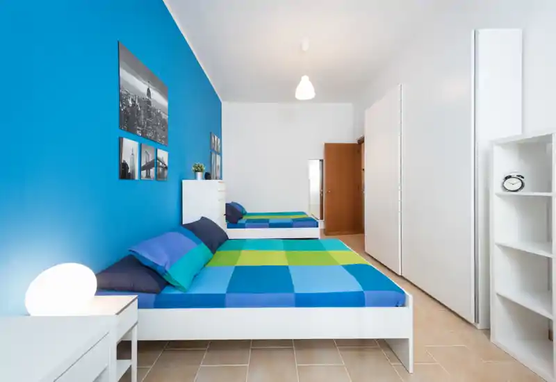 Camera da letto luminosa e moderna con due letti singoli con biancheria blu-verde coordinata, parete d'accento blu, mobili bianchi minimalisti e pavimento piastrellato. L'ambiente è fresco e ben illuminato, ideale per affitti brevi o camere per ospiti.