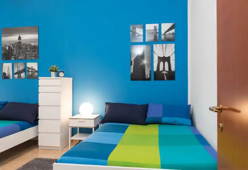 Camera da letto moderna e luminosa con biancheria colorata, comodino con lampada, cassettiera e quadri in bianco e nero su una parete accentuata blu intenso.