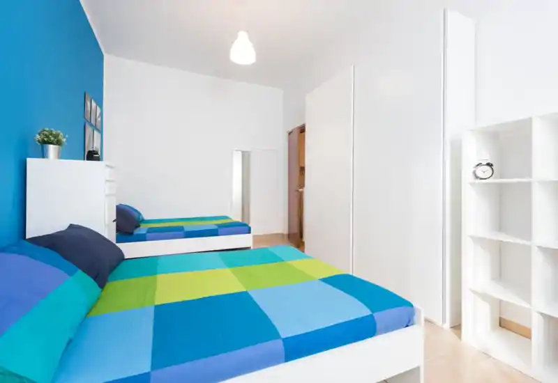 Camera da letto luminosa e moderna con due letti singoli con copriletti blu e verde, mobili bianchi minimal e una parete d'accento blu.