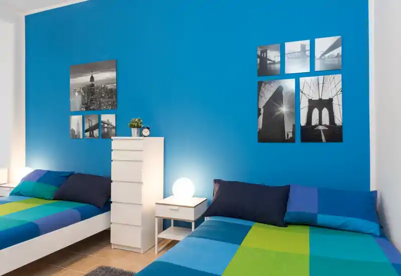 Chambre moderne avec deux lits simples, literie colorée et mur bleu vif décoré de photos en noir et blanc. Parfaite pour courts séjours ou colocataires, avec tables de chevet et éclairage doux pour une ambiance chaleureuse.