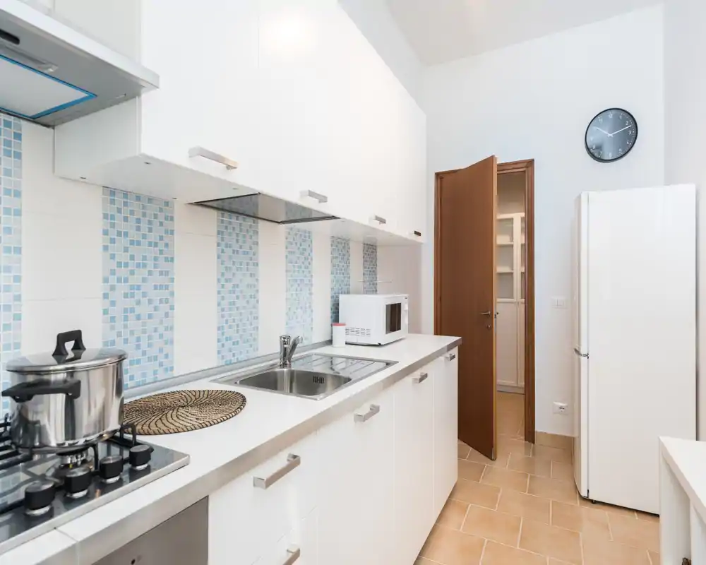 Cuisine moderne et lumineuse avec meubles blancs, crédence carrelée et évier inox; cuisinière, micro-ondes et réfrigérateur visibles—propre et bien agencée.