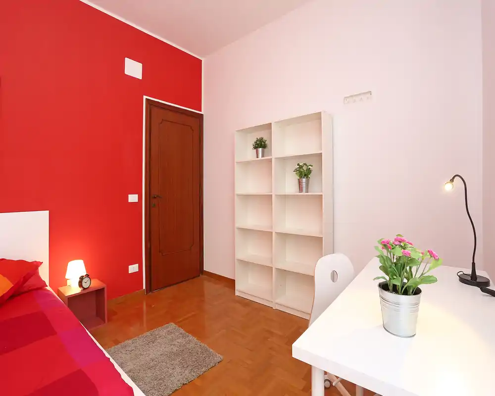 Chambre lumineuse et bien aménagée avec un mur rouge, un lit simple, une étagère et un bureau blanc avec des plantes — chaleureuse et moderne.