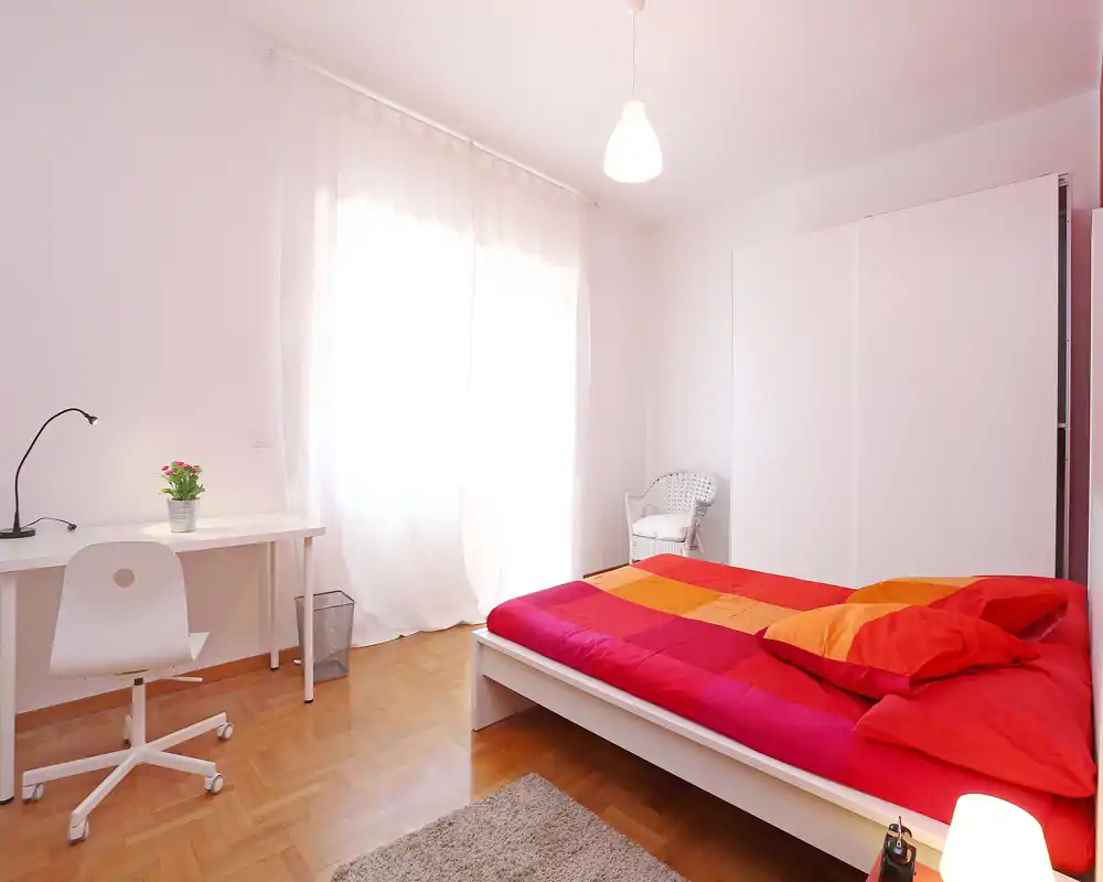 Chambre lumineuse et minimaliste avec lit double habillé de linge rouge et orange, armoire blanche, petit bureau et chaise près d'une grande fenêtre ensoleillée.