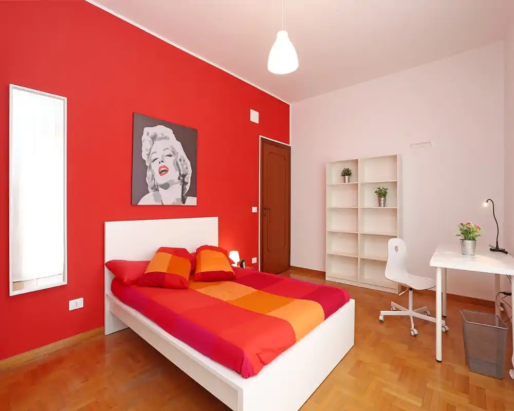 Chambre moderne et lumineuse avec lit blanc et couette rouge-orangée, parquet en bois, bureau et étagère — un espace chaleureux et bien éclairé, idéal pour les locations de courte durée.