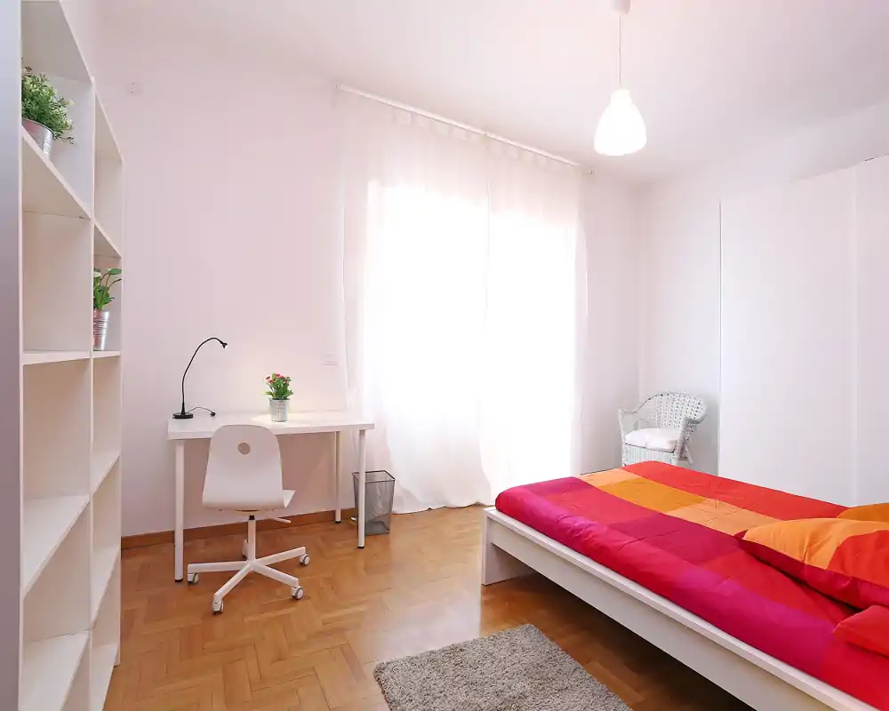 Chambre moderne et lumineuse avec un lit coloré, un bureau près d'une grande fenêtre et un mobilier minimaliste — idéale pour une annonce locative ou un séjour confortable.