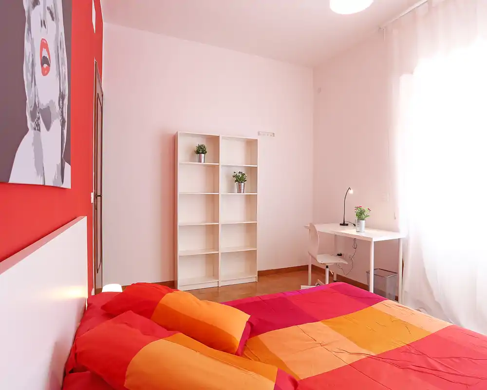 Chambre lumineuse et colorée avec un lit double au premier plan, étagère simple et petit bureau près de la fenêtre. La lumière naturelle et les textiles vifs donnent un espace agréable et bien mis en scène pour une annonce.