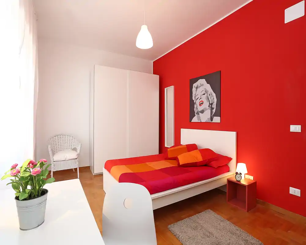 Chambre moderne et lumineuse avec un lit double habillé de couleurs chaudes rouges et orange, une armoire blanche, une table de chevet avec lampe et un tableau de Marilyn Monroe sur le mur rouge.