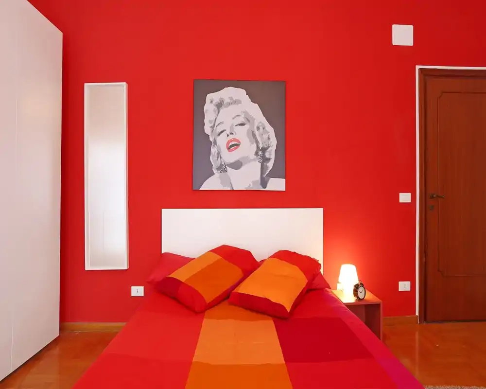 Chambre moderne et lumineuse avec un mur rouge vif, tête de lit blanche et linge de lit orange-rouge; lampe de chevet et affiche pop-art apportent chaleur et style.