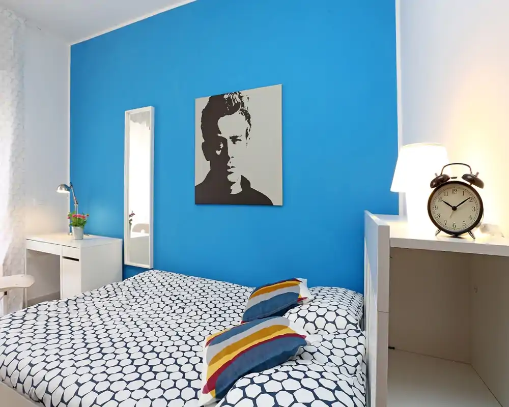 Dormitorio moderno y luminoso con pared azul intensa, ropa de cama estampada y cojines coloridos. Incluye mesilla con lámpara y despertador, escritorio pequeño y espejo de cuerpo entero — perfecto para alquileres de corta estancia.
