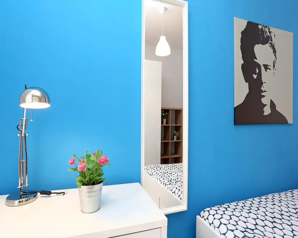 Rincón de dormitorio luminoso y moderno con pared azul, ropa de cama estampada, escritorio blanco con lámpara y maceta pequeña, y espejo de cuerpo entero que refleja parte de la habitación.