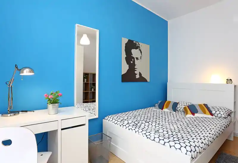 Dormitorio moderno y luminoso con cama blanca frente a una pared azul llamativa, ropa de cama estampada y un pequeño escritorio — bien presentado para anuncios.
