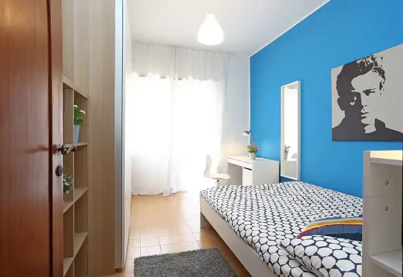 Dormitorio moderno y luminoso con cama doble de estampado, pared azul, escritorio y espejo junto a una gran ventana que aporta luz natural.