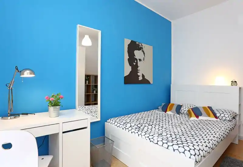 Bright, modern bedroom with a white bed and patterned bedding against a vivid blue accent wall; small desk and mirror make this a practical, well-staged room.