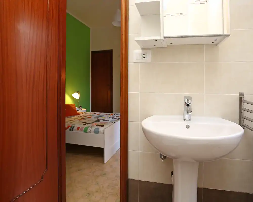 Dormitorio luminoso visto a través de una puerta con una cama de marco blanco, ropa de cama estampada y una lámpara de mesita — foto tomada desde el baño, muestra la relación entre estancias.
