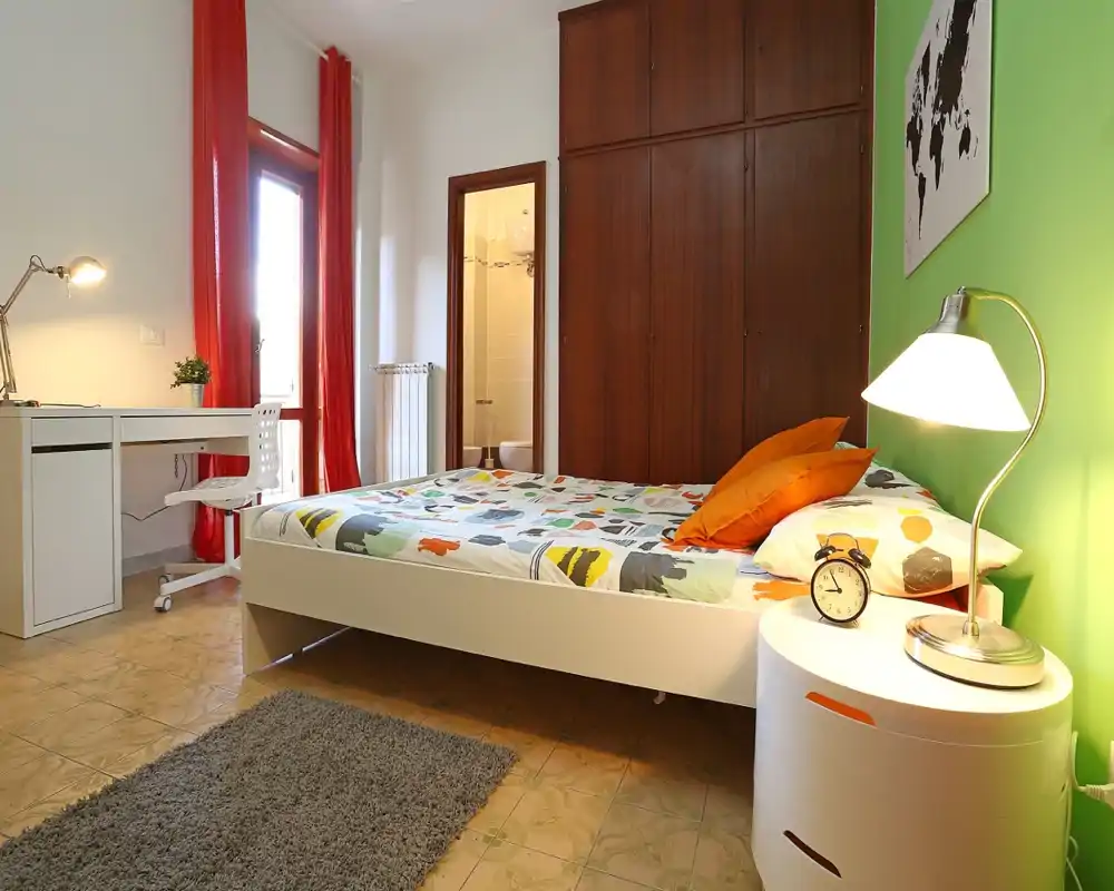 Dormitorio luminoso y bien decorado con cama doble, ropa de cama colorida y lámpara de mesita; escritorio junto a la ventana y armario empotrado.