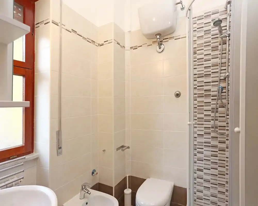 Baño compacto y bien iluminado con plato de ducha, inodoro, bidé y lavabo; azulejos beige neutros y accesorios modernos.