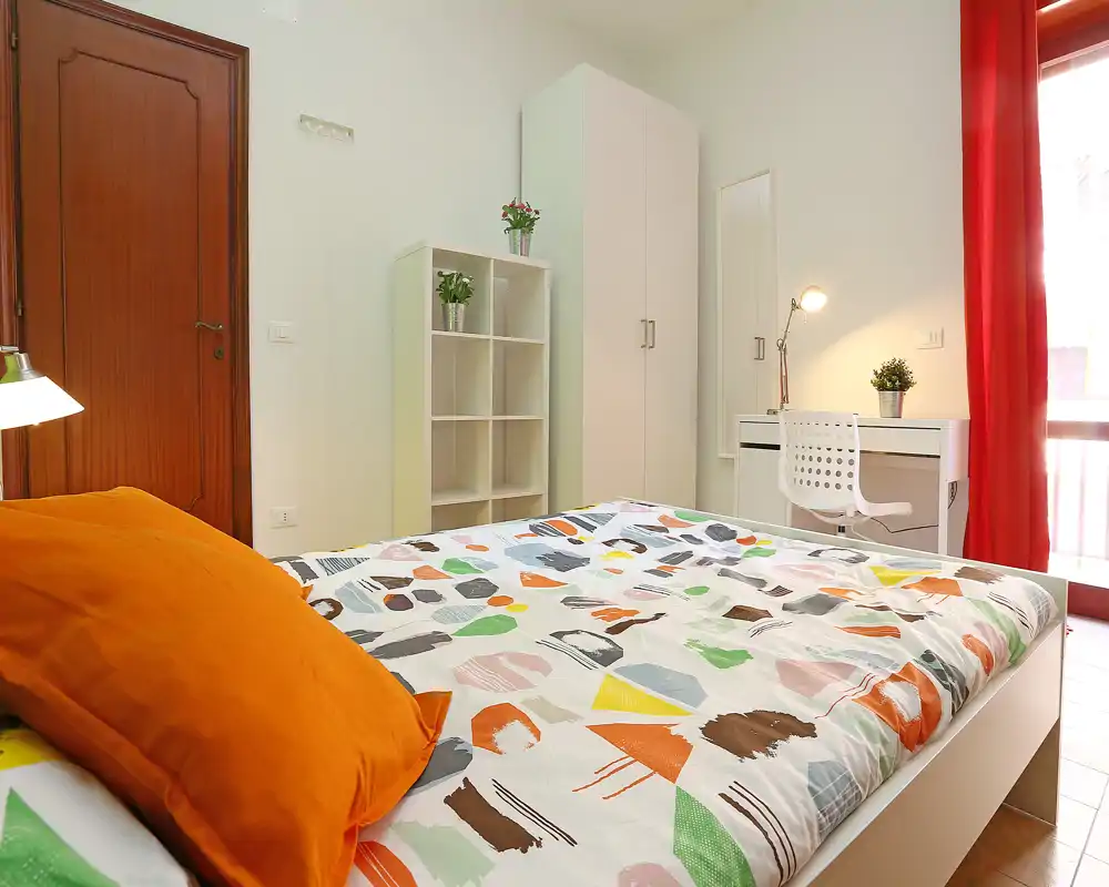 Dormitorio luminoso y bien presentado con ropa de cama colorida, cojín naranja, armario y un pequeño escritorio junto a la ventana que deja entrar luz natural.