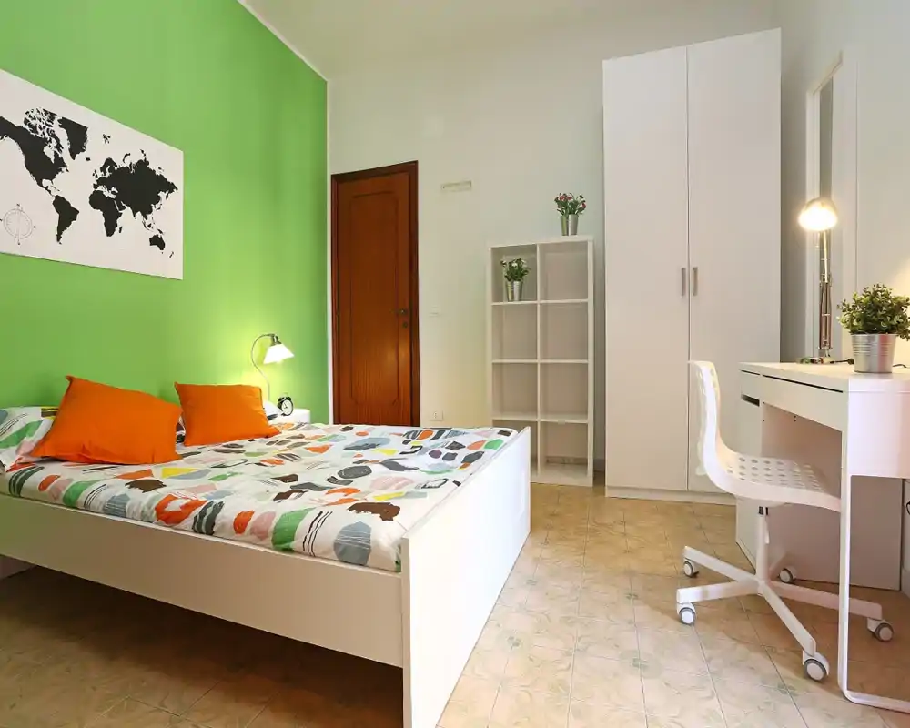 Dormitorio moderno y luminoso con una cama de estructura blanca, ropa de cama colorida y cojines naranjas frente a una pared verde brillante. La habitación incluye armario blanco, estantería y escritorio, ideal para estancias cómodas o alquileres de larga duración.