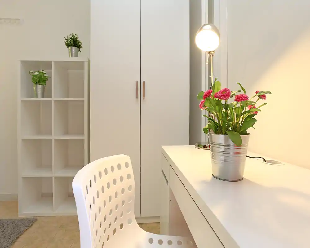 Rincón de dormitorio luminoso y minimalista con escritorio blanco, armario y plantas decorativas. La zona de trabajo ordenada y la luz suave crean una atmósfera tranquila y acogedora, ideal para leer o trabajar desde casa.