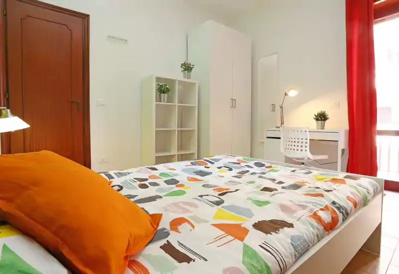 Dormitorio moderno y luminoso con edredón colorido, cojines naranjas, armario y escritorio junto a la ventana — ideal para un inquilino o un anuncio atractivo.
