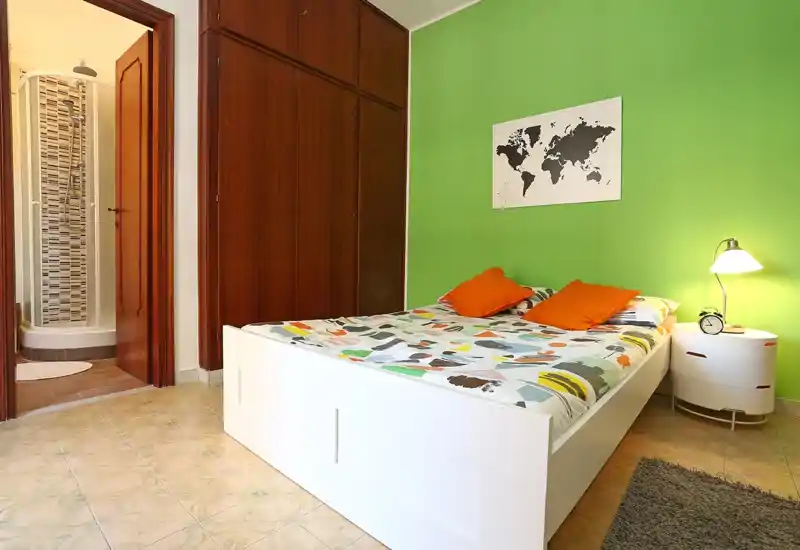 Dormitorio luminoso y bien presentado con cama blanca, ropa de cama estampada y dos cojines naranjas frente a una pared verde; armario empotrado de madera y puerta hacia el baño con ducha visibles.