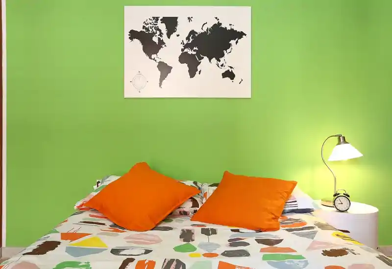 Dormitorio luminoso y moderno con edredón estampado, dos cojines naranjas y lámpara de mesita; un mapa del mundo en la pared verde aporta un toque vivo y con estilo.