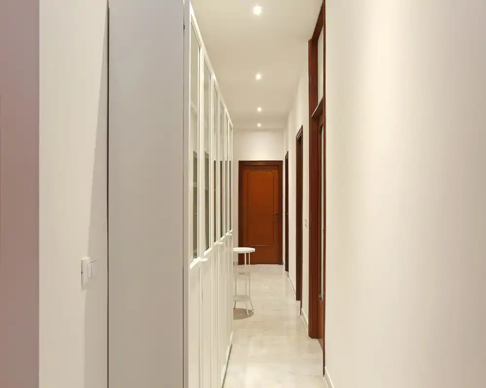 Pasillo de entrada luminoso y minimalista con focos empotrados, paredes blancas y puerta de madera al fondo — un recibidor limpio que conduce al interior.