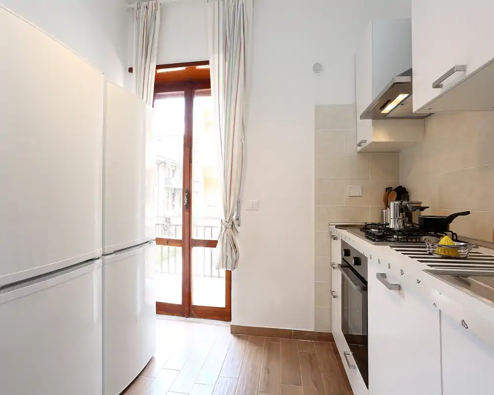 Cocina moderna y luminosa con armarios blancos, placa de gas, horno empotrado y frigorífico alto junto a una puerta de vidrio que da a un balcón. Suelo efecto madera y azulejos claros que crean un espacio acogedor.