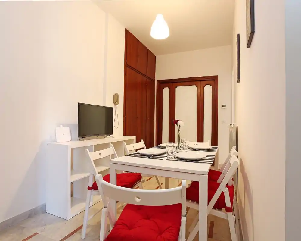 Área de comedor luminosa y ordenada con mesa blanca, sillas plegables con cojines rojos y un mueble TV sencillo; ideal para anuncios de pisos pequeños.