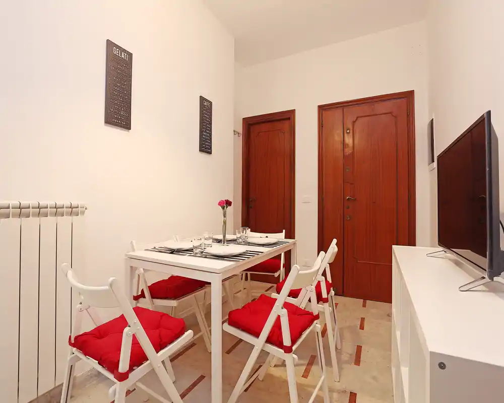 Zona de comedor compacta con mesa blanca para cuatro, sillas plegables con cojines rojos y un pequeño centro de mesa; espacio limpio, luminoso y bien preparado.