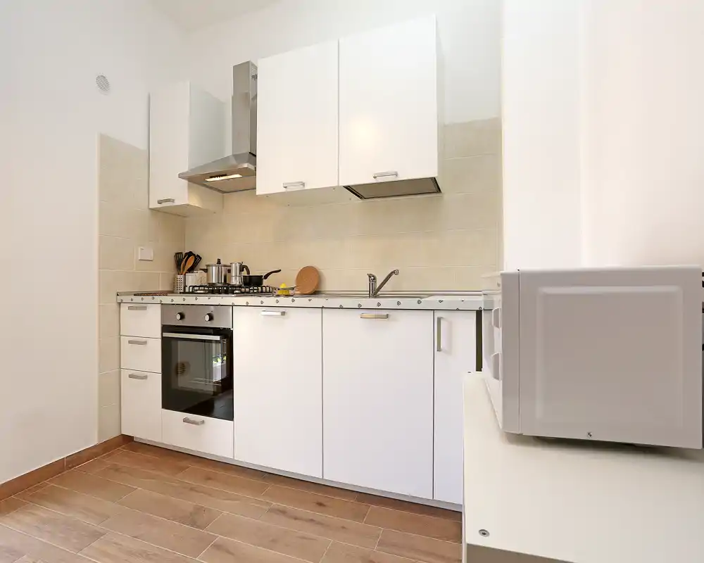 Cocina compacta y moderna, luminosa, con armarios blancos, horno integrado, placa de gas y campana de acero; suelo efecto madera y diseño limpio ideal para un apartamento funcional.