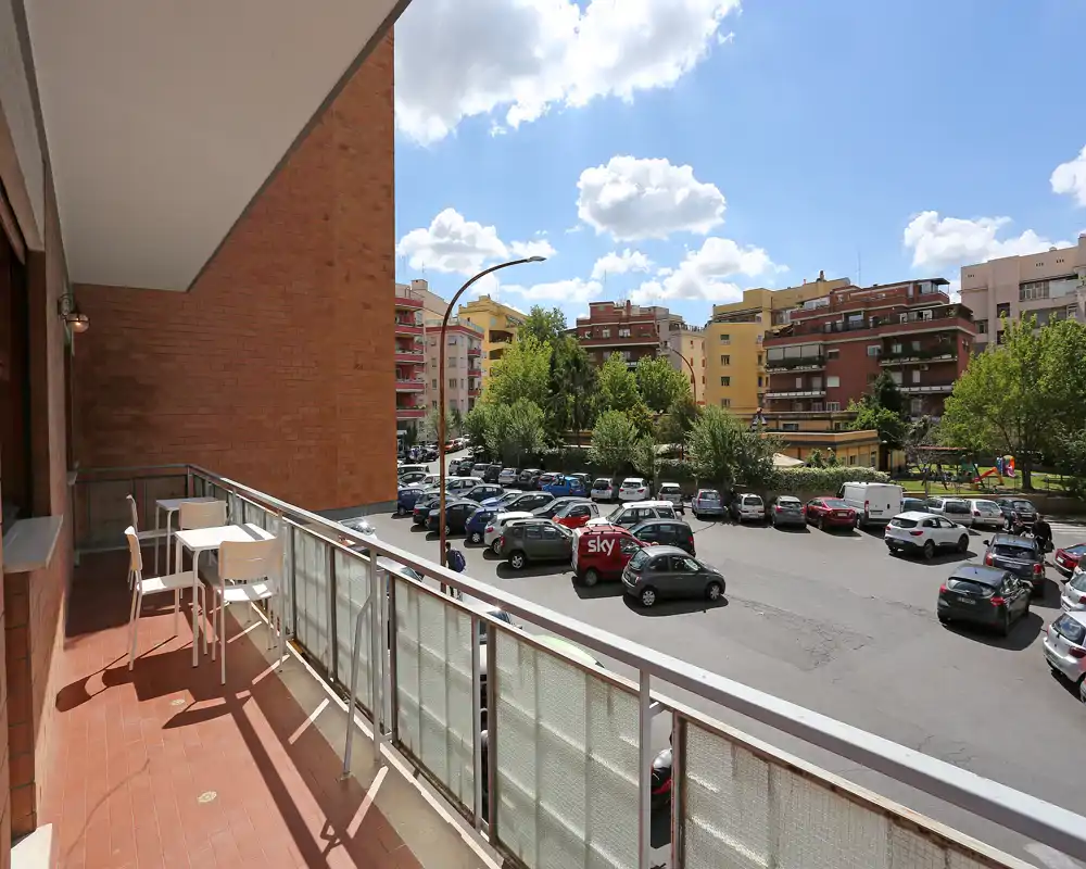 Balcón soleado con mesa y sillas, con vistas a un aparcamiento y edificios residenciales con árboles — espacio exterior luminoso y aireado.