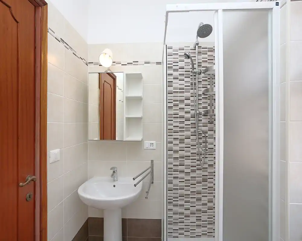 Baño luminoso y ordenado con lavabo de pie, armario con espejo y ducha acristalada con detalle de mosaico.