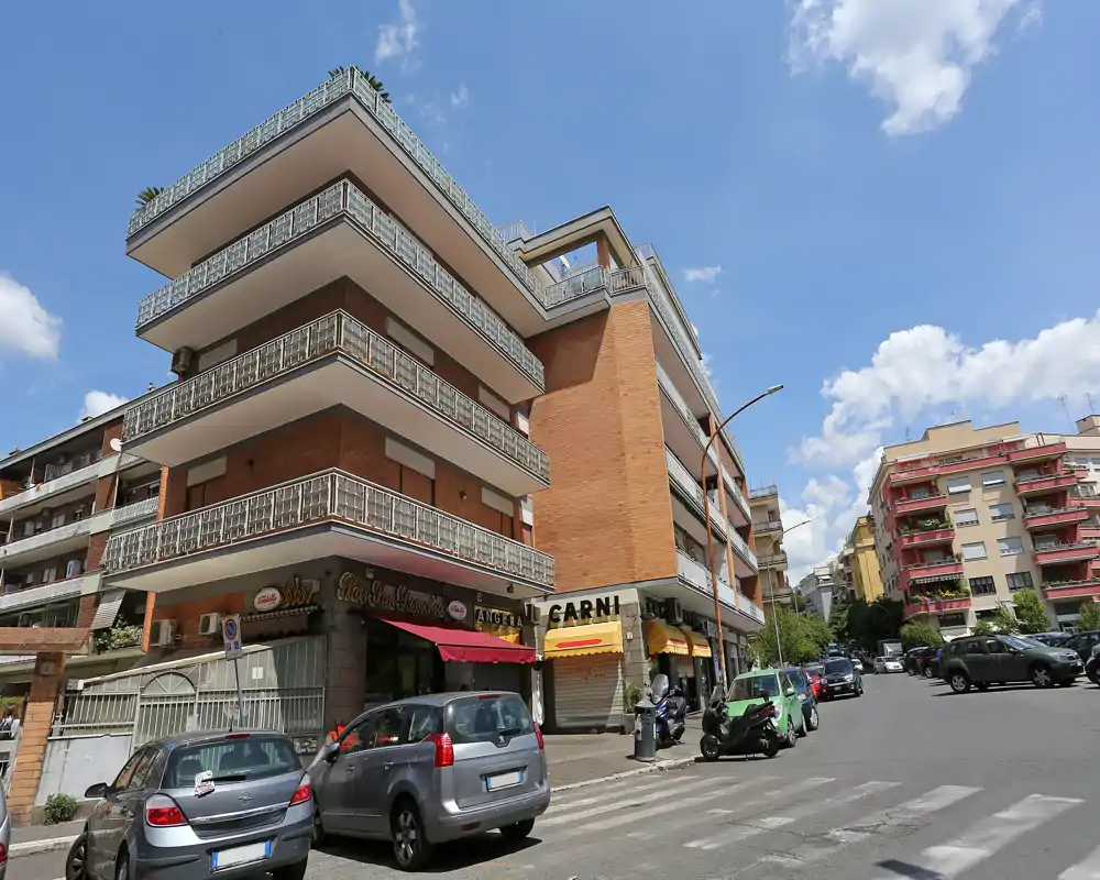 Vista exterior de un edificio residencial en la calle con amplios balcones, locales en planta baja y coches aparcados bajo un cielo azul.