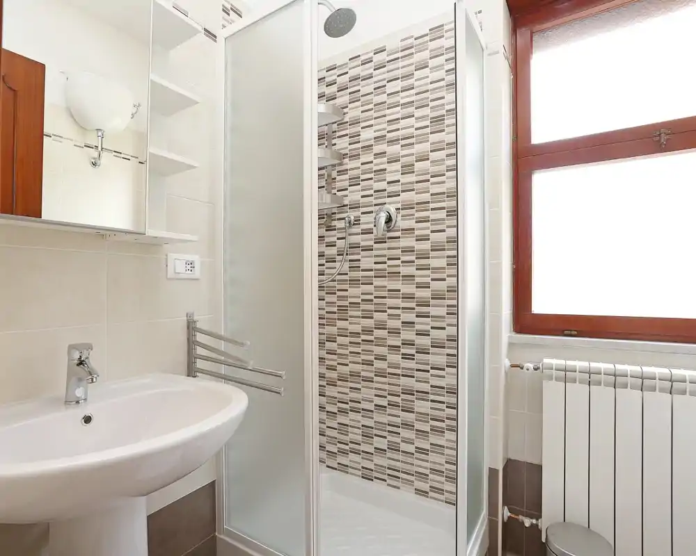 Baño limpio y bien iluminado con cabina de ducha en esquina con azulejos decorativos, lavabo de pedestal, espejo con armario y una gran ventana esmerilada que aporta luz natural.