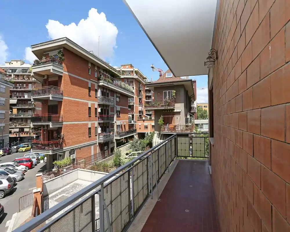Balcón privado soleado con vistas a edificios de ladrillo y un pequeño patio de aparcamiento; suelo de baldosas, barandilla metálica y espacio para sillas o plantas.
