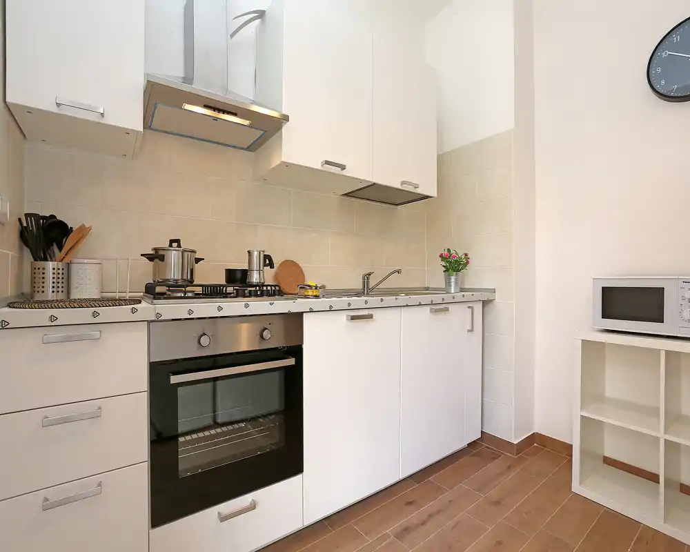 Cocina compacta y luminosa con armarios blancos, horno empotrado y placa de gas, microondas en una estantería y una pequeña planta decorativa: moderna y funcional.