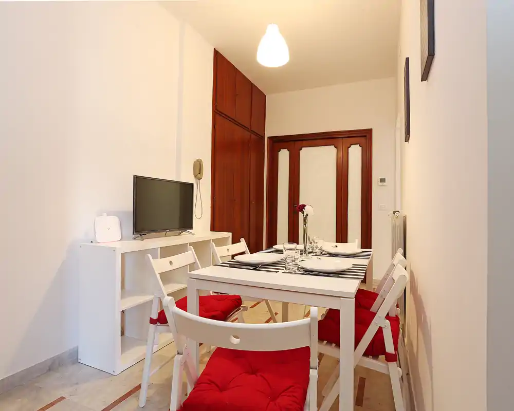 Zona de comedor luminosa y ordenada con mesa blanca, cojines rojos y una consola con TV — ideal para comidas en un apartamento pequeño.
